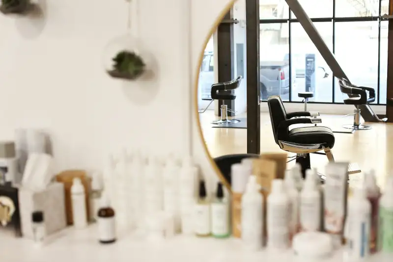 Runder Spiegel über einem Regal mit Pflegeprodukten, dahinter Friseurstühle vor großen Fenstern